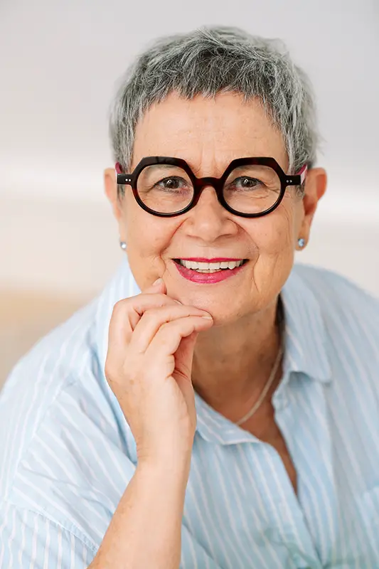 portrait-andrea_schweigler-physiopraxis-freiburg_9233b©ALEXJUNG Eine ältere Frau mit kurzem grauem Haar, Brille mit dunklem Rahmen und hellblau gestreiftem Hemd lächelt freundlich und stützt das Kinn auf die Hand.